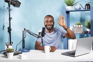 glimlachen blogger opname voor online kanaal, golvend Hoi naar abonnees en op zoek Bij camera. vrolijk Afrikaanse Amerikaans vlogger streaming leven terwijl werken in huis studio portret foto