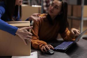 pakhuis werknemer vragen collega over plukken ticket Aan laptop terwijl werken Bij bureau in ecommerce kleinhandel bedrijf opslagruimte kamer. collega's bijhouden pakket Verzending en voorbereidingen treffen doos voor verzending foto