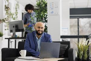 glimlachen bedrijf ondernemer werken Aan laptop en op zoek Bij camera in begin omhoog bedrijf kantoor. geslaagd project manager gebruik makend van portable computer terwijl zittend Aan bankstel portret foto