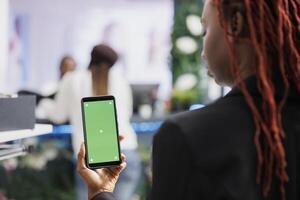 Afrikaanse Amerikaans vrouw gebruik makend van smartphone met groen scherm terwijl boodschappen doen voor kleren in winkelcentrum. winkel koper Holding mobiel telefoon met leeg chroma sleutel Scherm voor kopiëren ruimte foto