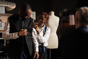 Afrikaanse Amerikaans naaister helpen cliënt proberen Aan kleermakers- stuk na afwerking handwerk. klant gelukkig met op maat gemaakt elegant kleding ontwerp in atelier winkel rennen door senior kleermaker foto
