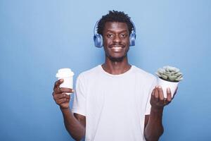 portret van gelukkig Afrikaanse Amerikaans Mens staand in voorkant van een blauw achtergrond met een ingemaakt fabriek terwijl draag- een drank. glimlachen zwart vent Holding een koffie kop en vervelend draadloze koptelefoon. foto