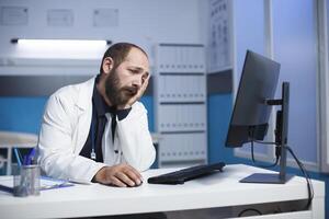 gebaard Kaukasisch Mens vervelend een laboratorium jas, vermoeid herzien medisch informatie Aan zijn bureaublad pc. mannetje dokter op zoek versleten uit is gebruik makend van computer in een ziekenhuis kantoor kamer. foto