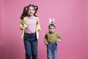 broer met konijn oren en zus jumping in de omgeving van in de studio, weinig kinderen wezen gelukkig over Pasen tijd viering. aanbiddelijk broers en zussen stuiteren Leuk vinden konijnen tegen roze achtergrond. foto