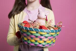 klein kind presenteren Pasen decoraties en speelgoed in een mand, Holding kleurrijk voorjaar feestelijk regelingen in voorkant van camera. jong positief meisje glimlachen in studio met aanbiddelijk decor. dichtbij omhoog. foto