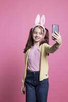jong opgewonden weinig kind nemen foto's met haar telefoon Aan camera, glimlachen in studio en poseren tegen roze achtergrond. positief blij kind duurt afbeeldingen voor Pasen vakantie evenement. foto