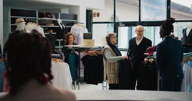 op te slaan werknemer helpen senior mensen met kleding suggesties, presenteren nieuw mode verzameling items naar paar in afdeling op te slaan. Afrikaanse Amerikaans werknemer maakt aanbevelingen. camera b. foto