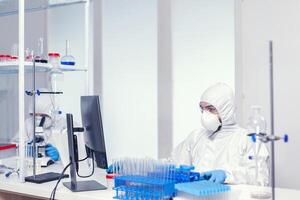 onderzoeker in steriel pak zittend Bij werkplaats in Onderzoek laboratorium voor covid19. medisch ingenieur gebruik makend van computer gedurende globaal pandemisch met coronavirus gekleed in overall. foto