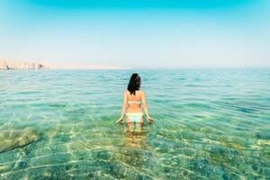 terug visie mooi toerist vrouw staan in Doorzichtig turkoois water in Perzisch golf mirella's eiland. Oman kustlijn paradijs. moesdam. foto