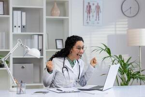 blij latino vrouw in medisch kleding juichen in voorkant van een laptop, uitdrukken succes en geluk in een kliniek instelling. foto