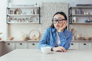 verdrietig en denken vrouw in keuken, depressief Aziatisch vrouw zittend Bij tafel Bij huis, jong vrouw in bril teleurgesteld. foto