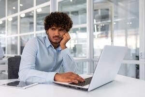 van streek jong zakenman werken binnen kantoor met laptop, Mens mislukt naar compleet technisch project Aan tijd, spaans Mens gefrustreerd zittend Bij bureau. foto