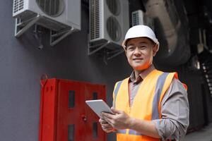 zelfverzekerd Aziatisch ingenieur in moeilijk hoed en reflecterende hesje glimlacht terwijl Holding een digitaal tablet, staand door hvac eenheden. foto