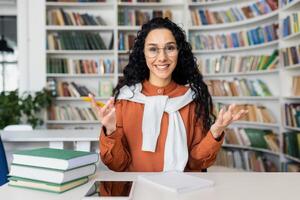 webcam visie online telefoongesprek, jong vrouw leerling pratend naar vrienden van een afstand, spaans vrouw binnen academisch bibliotheek Bij bureau aan het studeren en voorbereidingen treffen voor examens. foto