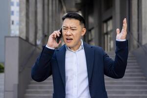 een van streek uitvoerend Aan een stressvol telefoon telefoongesprek, uitdrukken frustratie terwijl staand buiten kantoor gebouwen in een stedelijk instelling. foto