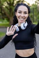 een Vrolijk, fit jong vrouw is gezien golvend Bij de camera, vervelend hoofdtelefoons in de omgeving van haar nek en training kleding, vertegenwoordigen een gezond en actief levensstijl. foto