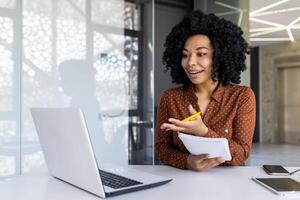 Afrikaanse Amerikaans vrouw met gekruld haar, vervelend een bruin blouse, actief bezighoudt met haar laptop terwijl Holding aantekeningen in een modern kantoor instelling. foto