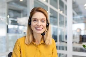 portret van volwassen vrouw klant onderhoud arbeider, volwassen bedrijf vrouw met telefoontje koptelefoon glimlachen en op zoek Bij camera, telefoontje centrum online klant onderhoud, binnen kantoor Bij werkplek. foto