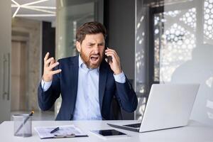 boos jong mannetje zakenman zittend Bij een bureau in de kantoor en pratend agressief Aan de telefoon, geschreeuw en gebaren met zijn handen. foto