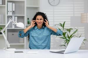 een professioneel vrouw in een blauw overhemd gevoel overweldigd door lawaai Bij haar kantoor bureau met een laptop, planten, en een lamp foto