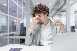 uitgeput volwassen zakenman tonen tekens van vermoeidheid en spanning terwijl werken Bij zijn kantoor bureau. wordt weergegeven hoofdpijn en nodig hebben voor een pauze. foto