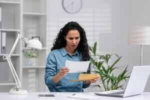 bezorgd latina zakenvrouw herzien een document met slecht nieuws in een modern huis kantoor instelling, uitdrukken spanning en frustratie. foto