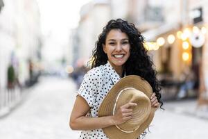 portret van een mooi speels Latijns Amerikaans vrouw, een jong toerist wandelen door de stad in de avond, glimlachen en op zoek Bij de camera, Holding een rietje hoed in haar hand. foto