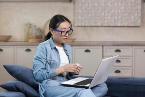 een jong mooi Aziatisch vrouw is zittend Bij huis Aan de bank, aan het kijken een film Aan een laptop, huilen, Holding een servet in haar handen. aangeraakt, besteedt de avond alleen. foto
