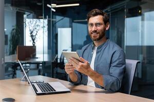 portret van een zakenman in een modern kantoor, een Mens toepassingen een tablet computer, zit Bij een bureau, glimlacht en looks Bij de camera in een gewoontjes overhemd en bril. foto