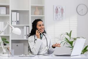 een vrolijk vrouw dokter met gekruld haar- gelukkig pratend Aan de telefoon terwijl op zoek Bij haar laptop in een goed verlicht, georganiseerd kantoor. foto