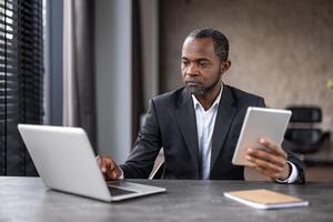 gefocust Afrikaanse Amerikaans zakenman toepassingen een laptop en tablet Bij zijn modern werkruimte, afbeelden toewijding en technologie gebruik in een professioneel instelling. foto