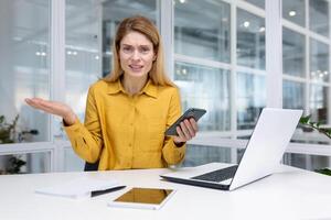 ontevreden blond vrouw zakenvrouw zittend Bij bureau in helder kantoor, Holding smartphone in hand, verliezende de loterij, lezing slecht nieuws, boos omdat van niet werkend telefoon, op zoek Bij de camera. foto