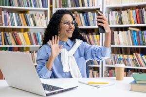 een blij vrouw in een bibliotheek bezighoudt in een telefoontje gebruik makend van haar smartphone, glimlachen en zwaaien, omringd door boeken en een laptop. foto