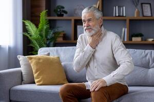ouderen heer met een baard gevoel ongemak in zijn keel, zittend Aan een sofa in een goed versierd leven kamer, uitdrukken zorg. foto