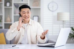 volwassen Aziatisch Mens pratend Aan de telefoon Bij huis, Mens werken in huis kantoor zittend Bij bureau in huis kantoor, gelukkig en lachend, overleg plegen klanten van een afstand. foto