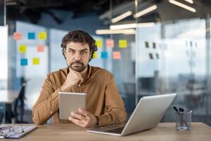 gefocust professioneel gebruik makend van een laptop en een tablet Bij een houten bureau in een modern kantoor, met kleverig aantekeningen in de achtergrond. foto