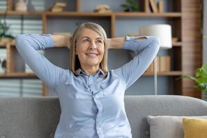 een sereen ouder vrouw met een inhoud glimlach, zit terug met haar handen achter haar hoofd in een modern leven kamer. ze straalt uit een kalm, ontspannen atmosfeer. foto