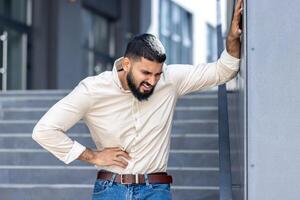 een jong moslim Mens in een overhemd is staand buiten een kantoor gebouw, leunend zijn hand- Aan de muur en Holding de kant van zijn lichaam, gevoel erge, ernstige pijn in zijn maag, grimassen van stuiptrekkingen foto