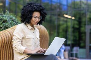 jong mooi zakenvrouw zittend Aan bank buiten kantoor gebouw, vrouw arbeider met laptop werken buitenshuis, Afrikaanse Amerikaans vrouw werken online van een afstand. foto