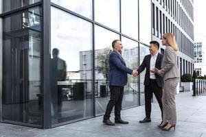 vergadering van drie geslaagd bedrijf mensen, verschillend droom team Mens en vrouw buiten kantoor gebouw, groet en beven handen, ervaren professionals specialisten in bedrijf pakken pratend foto