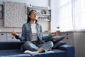 mooi Aziatisch vrouw ontspannende Bij huis zittend Aan sofa en mediteren in lotus houding, aan het doen ademen opdrachten in leven kamer foto