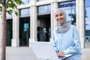 glimlachen professioneel vrouw vervelend een hijab werken Aan haar laptop, gezeten buiten een kantoor gebouw met een modern facade. foto