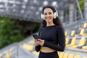 een jong, fit vrouw met een smartphone en hoofdtelefoons staat glimlachen Bij een sport- stadion, uitstralend energie en welzijn. foto