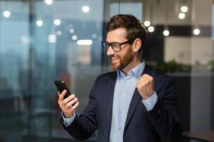 geslaagd zakenman investeerder gebruik makend van telefoon, Mens vieren zege en geslaagd prestatie, Holding hand- omhoog triomf gebaar, baas in bedrijf pak binnen kantoor staand in de buurt venster. foto