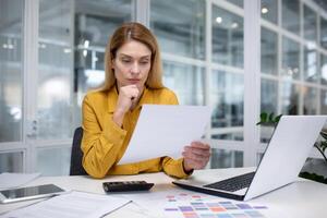 van streek blond zakenvrouw werken in een modern kantoor, zittend Bij een bureau in voorkant van een notitieboekje, bedachtzaam werken met bedrijf papieren, voorzichtig herzien belangrijk informatie. foto