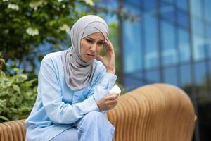 een middelbare leeftijd vrouw vervelend een blauw hijab looks verontrust en onwel, zittend buitenshuis met weefsels in haar hand, uitdrukken ongemak of ziekte in een stedelijk instelling. foto