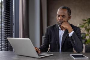 attent mannetje uitvoerend in pak verloofd in diep overpeinzing Bij zijn kantoor bureau met laptop en digitaal tablet. foto