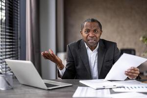 perplex Afrikaanse zakenman gevoel gefrustreerd terwijl Holding documenten en werken Aan een laptop Bij zijn kantoor bureau. foto