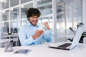 een jong Indisch zakenman is werken in de kantoor Bij een tafel met een laptop, Holding een telefoon in zijn handen. hij looks Bij de mobiel scherm, verheugt zich, viert succes, shows een zege gebaar met zijn hand. foto
