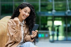 jong vrouw in gewoontjes bedrijf kleding verdiept in haar smartphone terwijl gezeten buitenshuis met een modern gebouw in de achtergrond, symboliseert connectiviteit en stedelijk levensstijl. foto